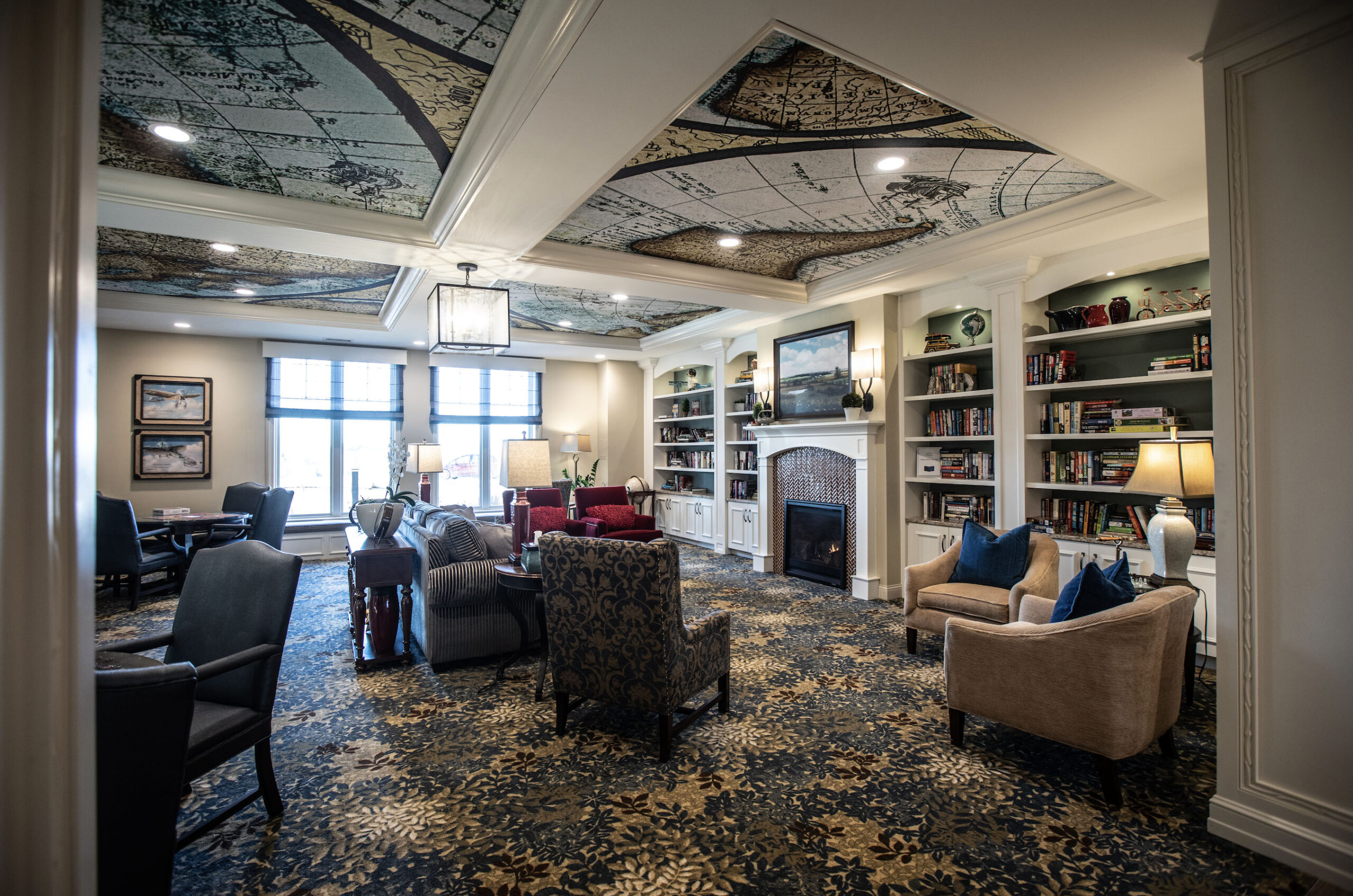 Rivertown Ridge community space with warm carpeted flooring, bright windows, and a wall of books with a fireplace and comfortable furniture.