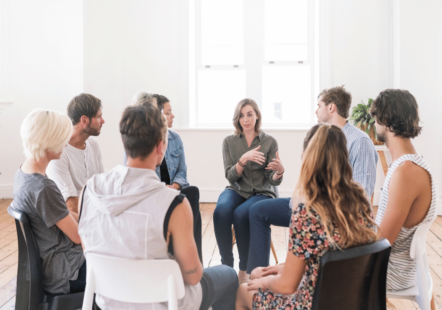 Group of adults seated in a circle in a bright room, woman in center speaking with hand gestures while others listen.